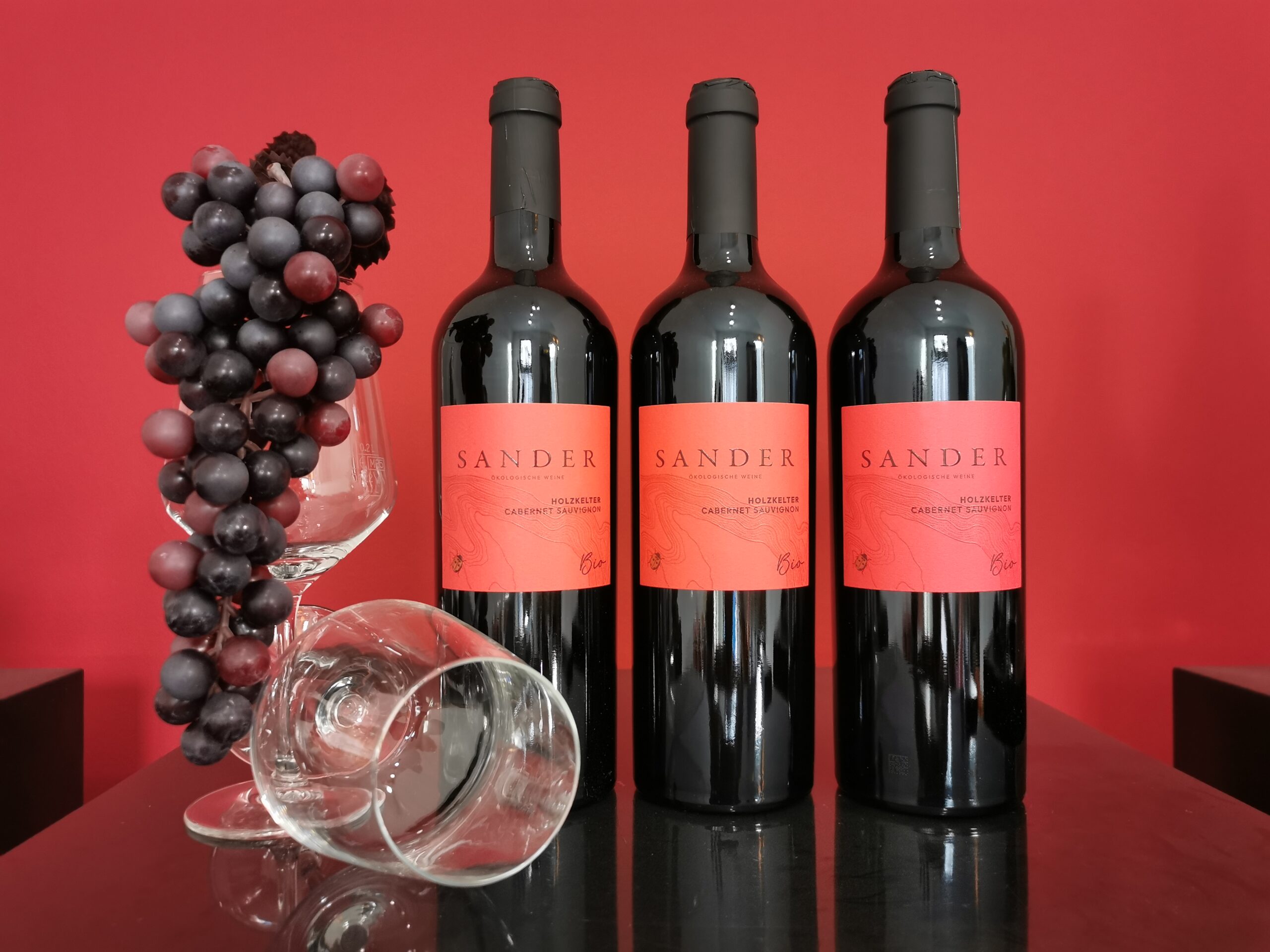 Flasche SANDER Cabernet Sauvignon Holzkelter Bio auf schwarzem Untergrund mit Weinglas und Trauben.