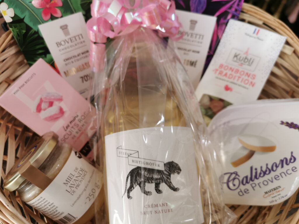 Ein liebevoll arrangierter Gourmet‑Geschenkkorb mit französischen Delikatessen: Crémant‑Flasche, Lavendelhonig, Schokolade, Calissons, Bonbons und rosa Biscuits. Warmes Licht, natürliche Materialien und ein festlicher Ostercharakter.